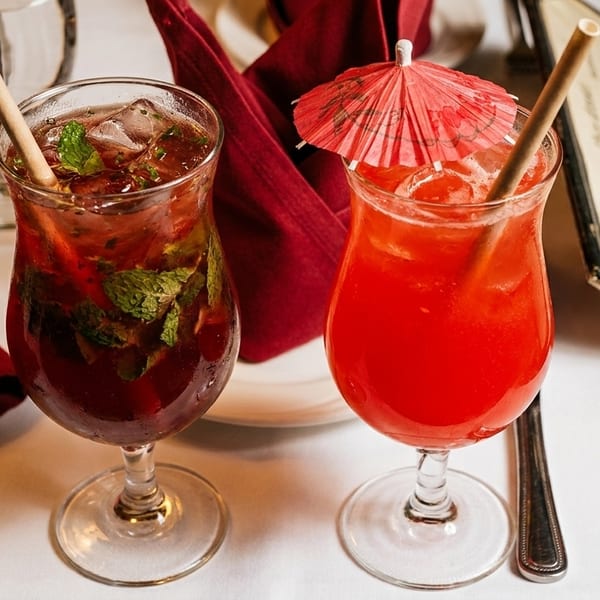 Two vibrant cocktails, one red with a paper umbrella, on a white tablecloth with a menu.
