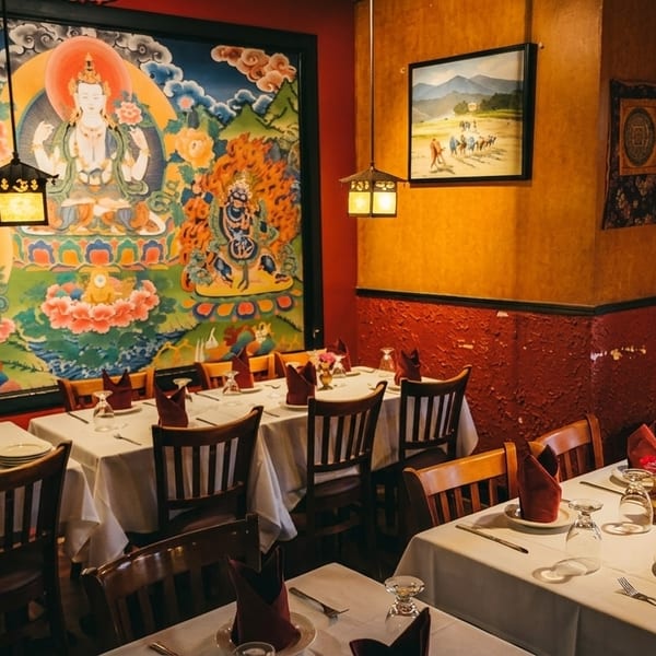 Restaurant dining room with white-clothed tables, wooden chairs, and a large colorful mural on the wall.