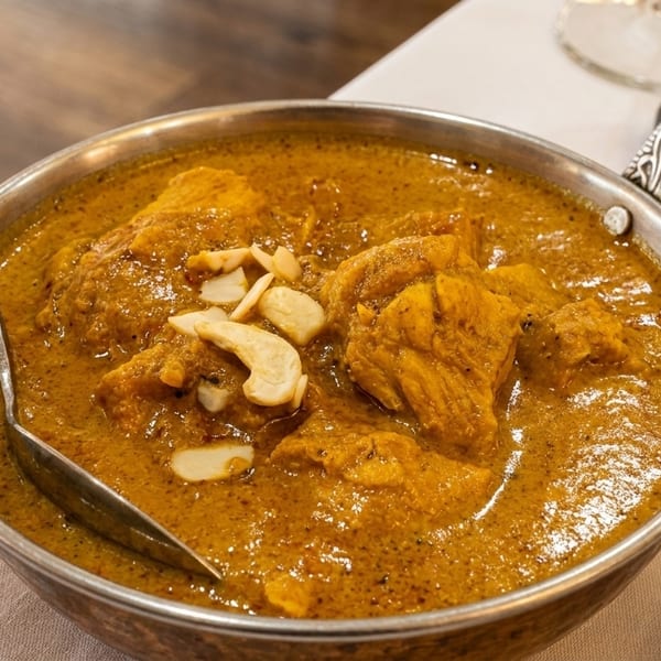 A metal bowl of chicken curry with cashews and a spoon, on a white tablecloth.