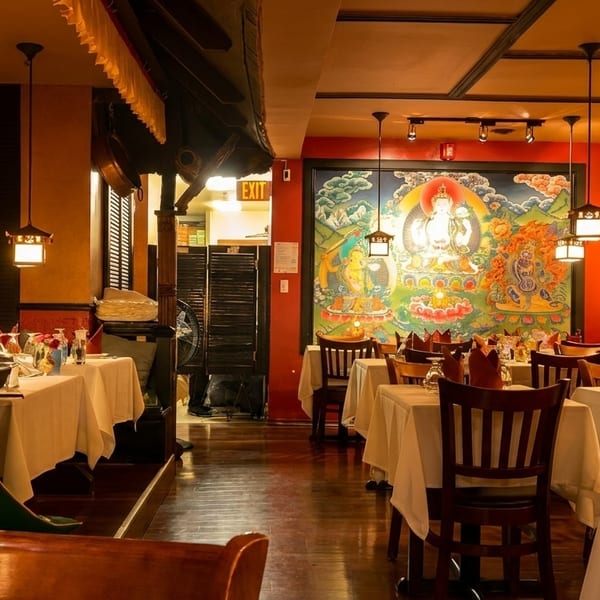 Warmly lit restaurant interior with white-clothed tables, wooden chairs, and colorful Buddhist thangka paintings on red walls.