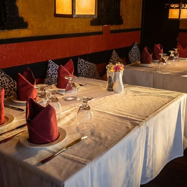 Restaurant dining room with white tablecloths, red napkins, and patterned pillows on banquette seating.